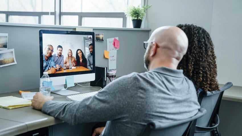 a man and a woman in a cubicle looking at a monitor where they are holding a video conference call with another professional team